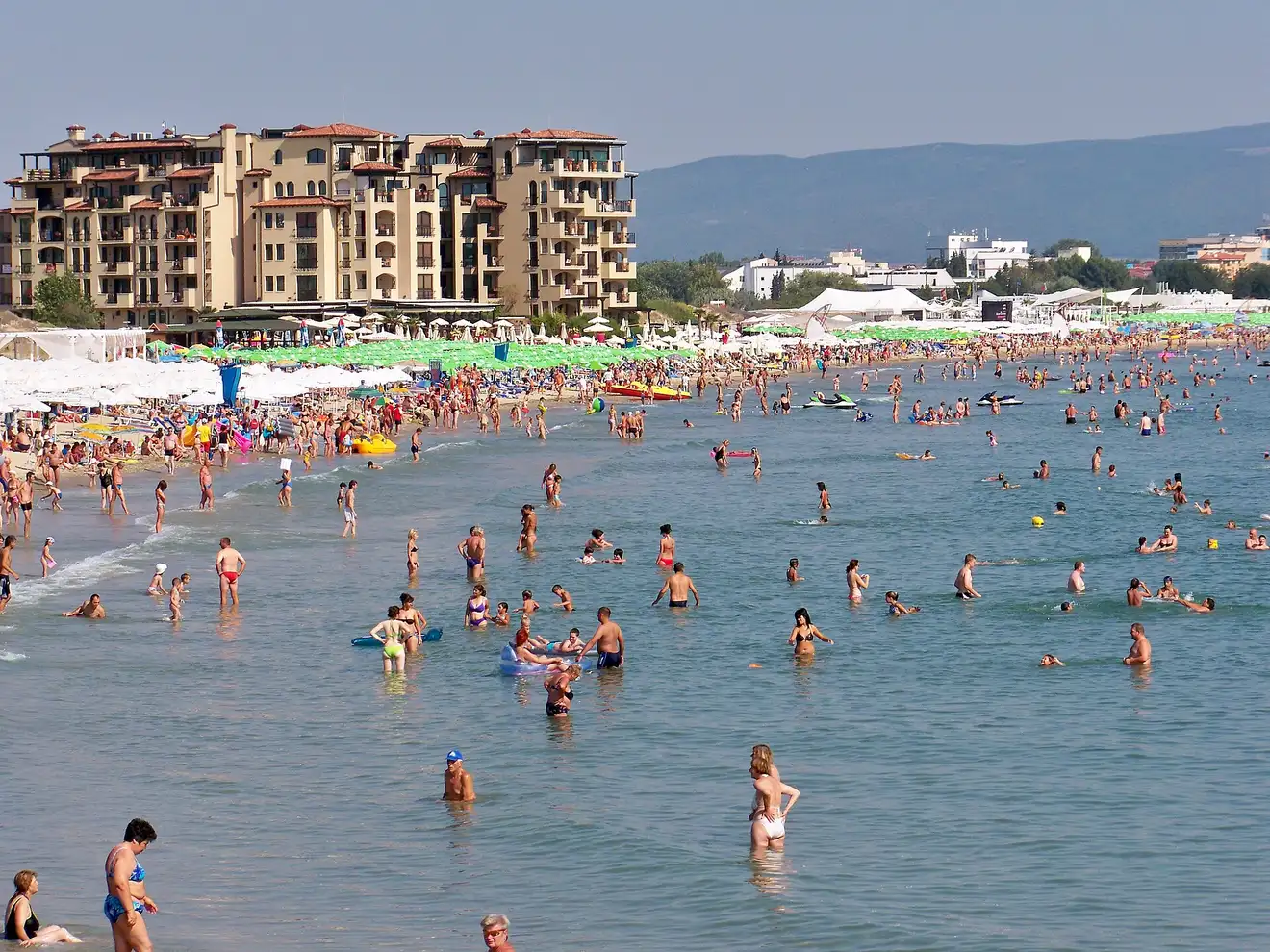 The packed beach at Sunny Beach with crowds swimming in the shallow sea, rows of umbrellas and a wall of resort hotels with forested hills behind