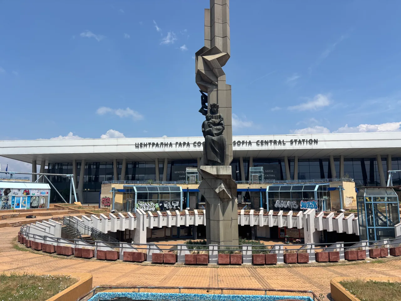 The forecourt of Sofia Central Station with a concrete monument, graffiti on the lower shopfronts and the station sign behind
