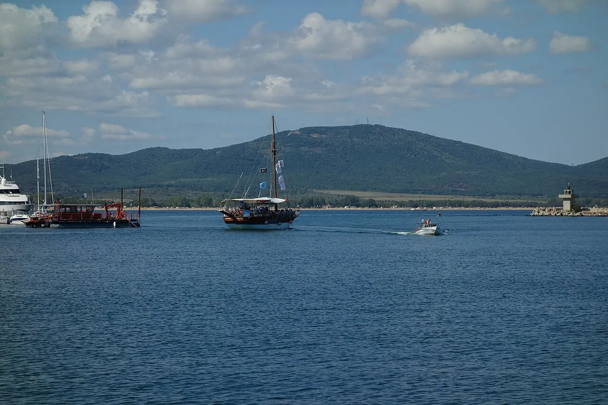 A pleasure boat leaving Sozopol harbour past the breakwater lighthouse, with the marina and green hills behind