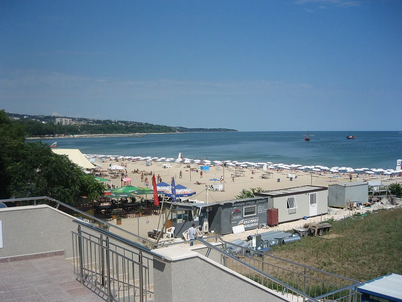 Varna's central city beach in summer, rows of white parasols and sunbeds on golden sand, the bay curving away to a green headland with boats offshore