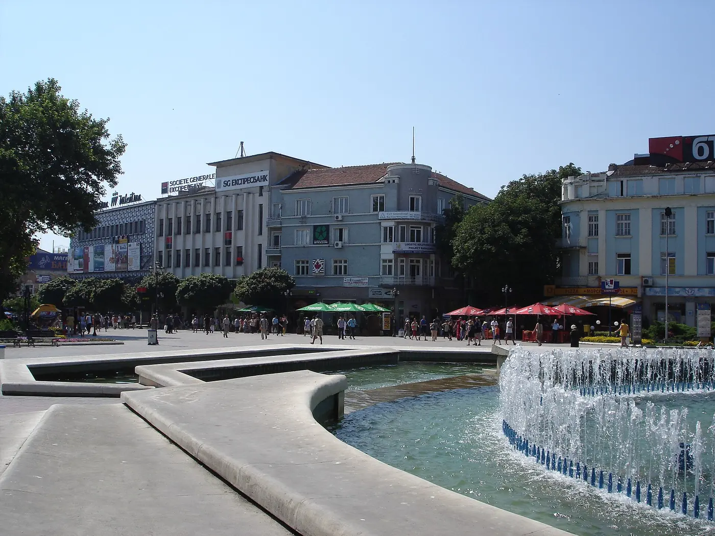 The central pedestrian square in Varna with a fountain, cafes with green and red umbrellas, and people strolling past early-1900s facades