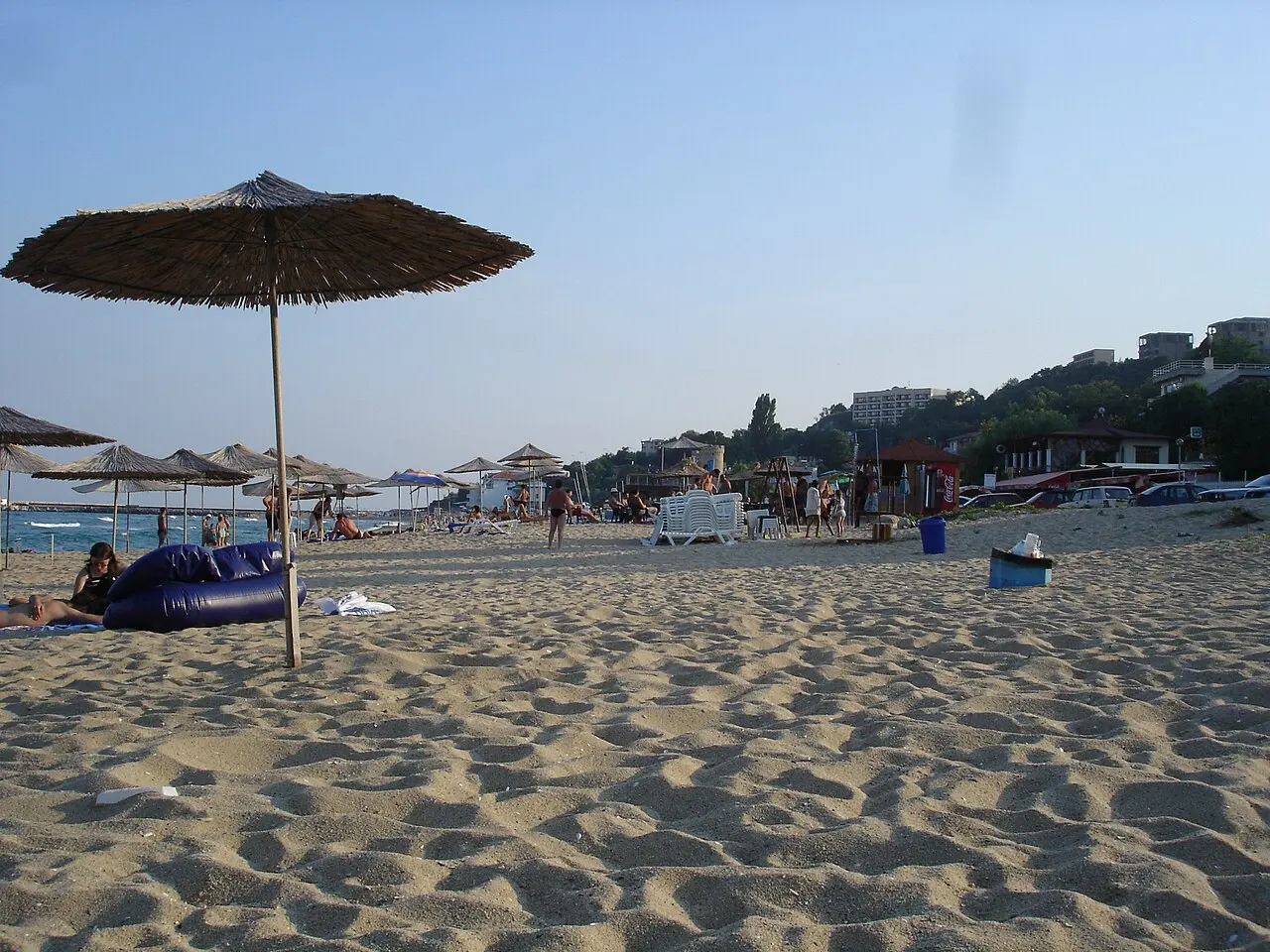 The sandy Kabakum beach north of Varna in the late afternoon, thatched straw parasols on the sand, swimmers in the sea and low green hills with hotels behind