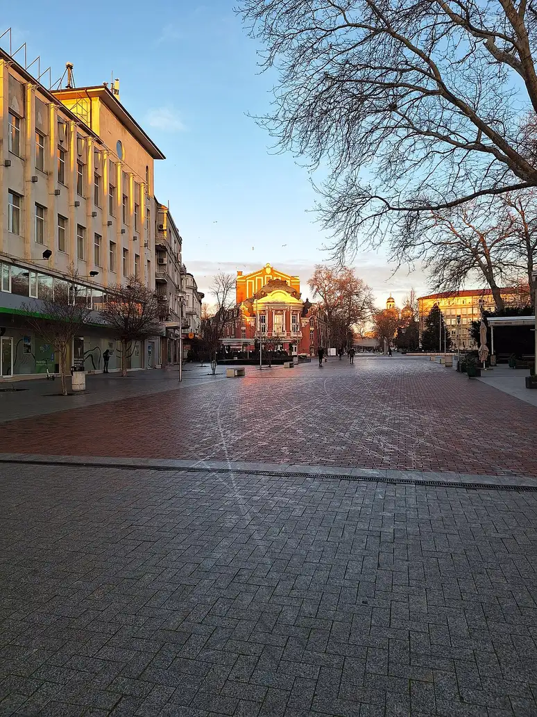 The pedestrian Nezavisimost Square in central Varna at golden hour, its brick paving empty, with the ochre Varna Opera building lit at the far end