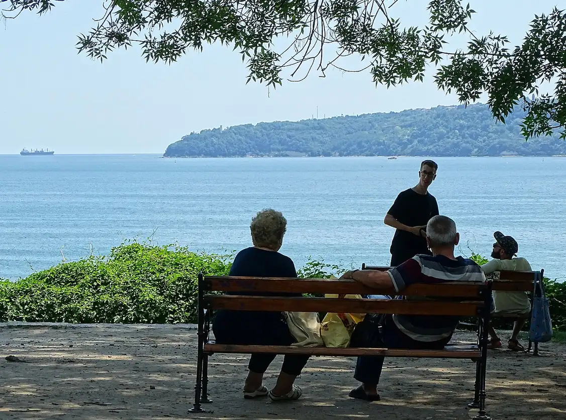 People sitting on a bench in the shade of trees in Varna's Sea Garden, looking out over the calm blue Black Sea to a green headland across the bay